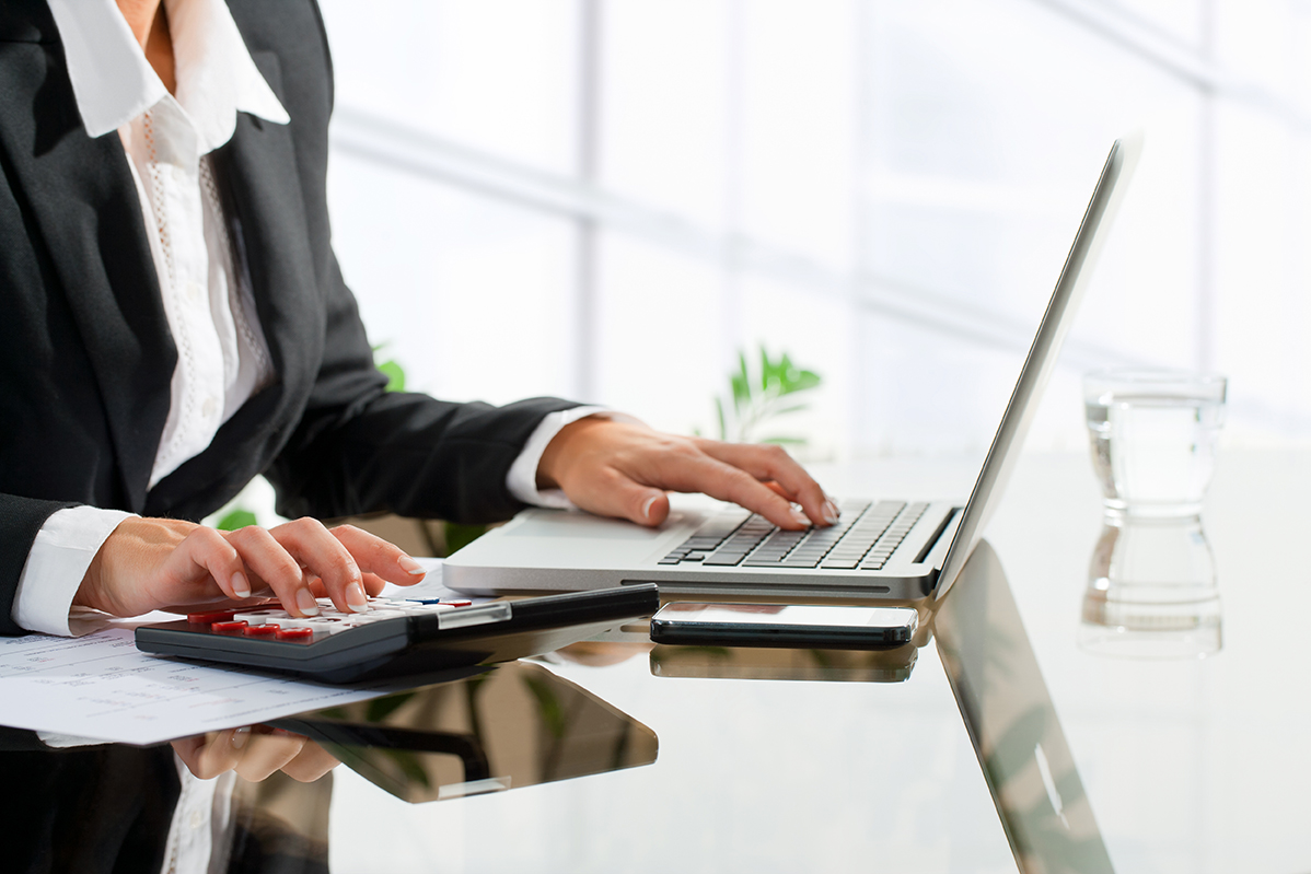 Close up of female office worker's hands doing accounting with calculator and laptop.